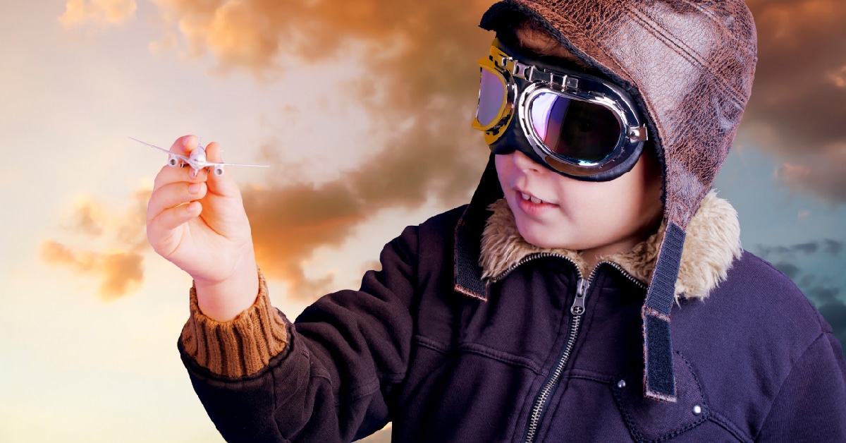 Boy in a pilot helmet and aviator goggles flies a toy plane.